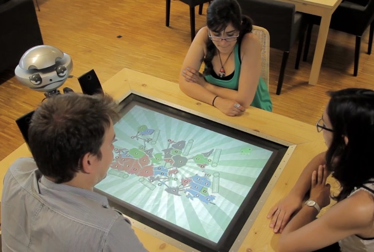 Three players gathered around the digital multi-touch Risk board game, with the EMYS robot at the table